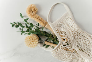 Two natural fiber dish brushes with wooden handles placed in a cotton mesh bag alongside eucalyptus sprigs, arranged on a light surface for a clean, eco-friendly aesthetic.