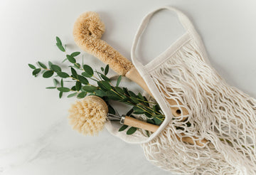 Two natural fiber dish brushes with wooden handles placed in a cotton mesh bag alongside eucalyptus sprigs, arranged on a light surface for a clean, eco-friendly aesthetic.