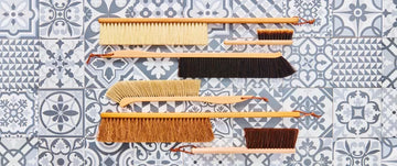 Assorted natural fiber brushes with wooden handles displayed on patterned blue and white tile background.
