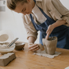 Female artisan shaping a clay vessel by hand on a wooden work table, surrounded by pottery tools and slip bowls in a sunlit ceramics studio.