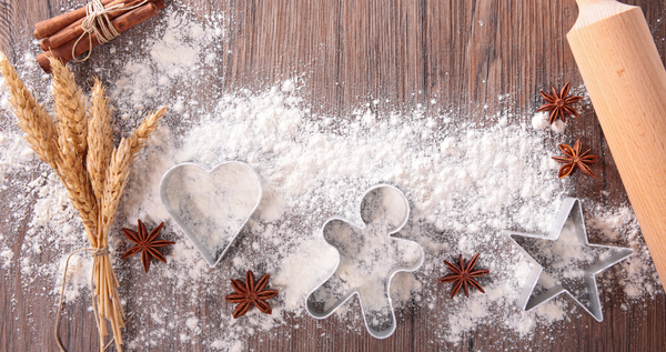 Holiday baking flat lay with flour-dusted cookie cutters (heart, gingerbread, star), rolling pin, wheat stalks, and star anise on a wooden surface.