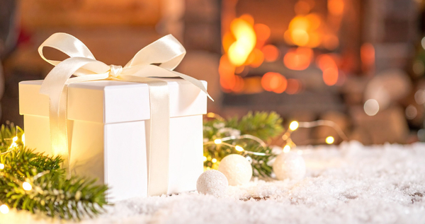White gift box with satin ribbon on snowy greenery in front of a warm fireplace—holiday scene.