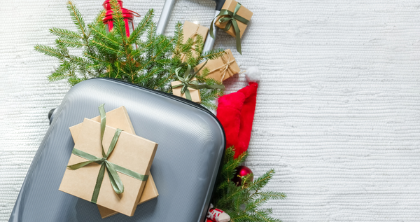 Carry-on suitcase with small wrapped gifts, fir greenery, and a Santa hat—festive holiday travel.