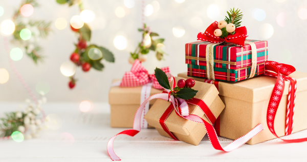 Stacked holiday gift boxes with red ribbons and plaid wrap on a white table, with soft bokeh lights and greenery in the background.