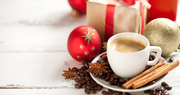 White espresso cup with cinnamon sticks, coffee beans, and star anise; red and gold ornaments and a wrapped gift in soft holiday light.